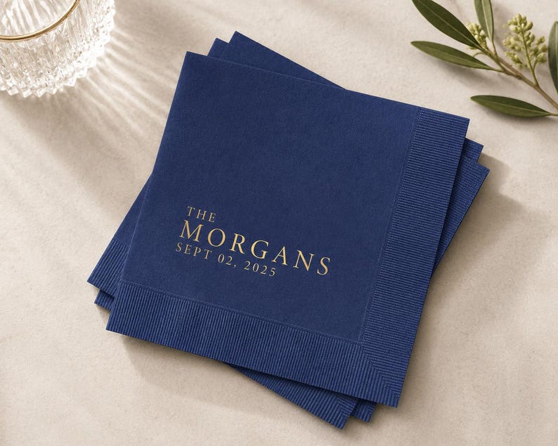 May include: A stack of square navy blue paper napkins with a textured border. The top napkin features gold foil text that reads THE MORGANS and SEPT 02, 2025, centered in a serif font. The napkins are layered slightly offset from one another.