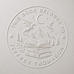 May include: A circular embossed stamp design featuring an open book resting on a stack of books, mountains, pine trees, two dragons, and a crescent moon. The text reads THIS BOOK BELONGS TO JEFFREY THOMASON in a circular border on a white background.