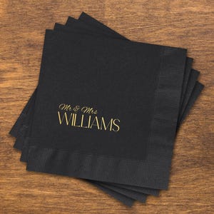 May include: A stack of square black paper napkins with a textured border. The top napkin features the text "Mr & Mrs" in a gold script font above the word "WILLIAMS" in a larger, gold, sans-serif capital letter typeface.