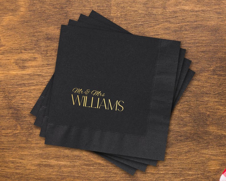 May include: A stack of square black paper napkins with a textured border. The top napkin features the text "Mr & Mrs" in a gold script font above the word "WILLIAMS" in a larger, gold, sans-serif capital letter typeface.