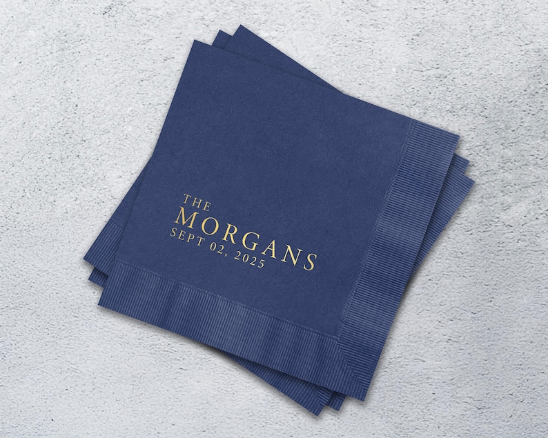 May include: A stack of square navy blue paper napkins with a textured border. The center features gold foil text that reads THE MORGANS and SEPT 02, 2025 in a clean, serif font. The napkins are layered slightly offset to show the edges.
