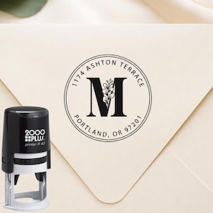 May include: A black and white 2000 Plus Printer R 40 self-inking stamp next to an envelope. The stamp impression features a circular border, the text 1174 ASHTON TERRACE PORTLAND, OR 97201, and a large letter M with a small leaf sprig design.