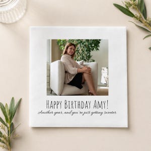 May include: A square white greeting card featuring a centered photograph of a person sitting in a white armchair. Below the photo, the text reads: Happy Birthday Amy! Another year, and you're just getting sweeter. The card has a clean, minimalist design.