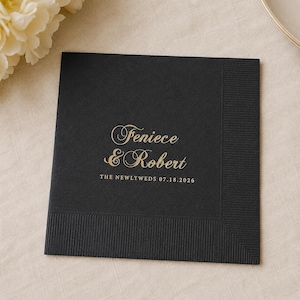May include: A square black paper napkin with a textured border. The center features gold foil lettering that reads Feniece & Robert, followed by the text THE NEWLYWEDS 07.18.2026 in a smaller, clean sans-serif font centered below the names.