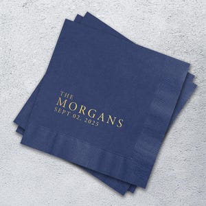 May include: A stack of square navy blue paper napkins with a textured border. The center features gold foil text that reads THE MORGANS and SEPT 02, 2025 in a clean, serif font. The napkins are layered slightly offset to show the edges.