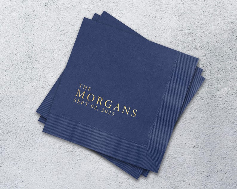 May include: A stack of square navy blue paper napkins with a textured border. The center features gold foil text that reads THE MORGANS and SEPT 02, 2025 in a clean, serif font. The napkins are layered slightly offset to show the edges.