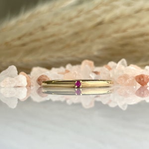May include: A thin gold band ring with a single small ruby gemstone set in the centre.