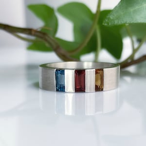 May include: A silver band ring with three rectangular gemstones. The gemstones are blue, red, and orange.