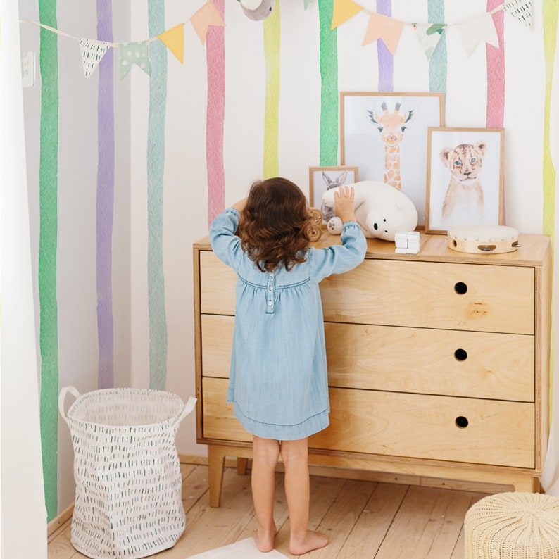 K&ouml;nnte beinhalten: Ein junges M&auml;dchen steht vor einer Kommode mit drei Schubladen. Die Kommode ist aus hellem Holz und hat ein schlichtes Design. Das M&auml;dchen tr&auml;gt ein blaues Jeanskleid und hat lange braune Haare. Die Wand hinter der Kommode ist mit bunten Streifen bemalt. Auf der Kommode stehen mehrere gerahmte Bilder, darunter eines mit einer Giraffe und eines mit einem L&ouml;wen.