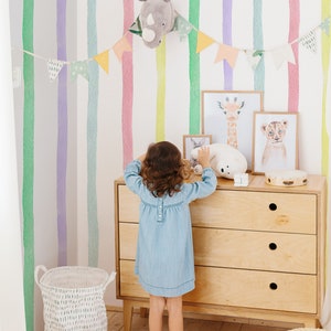 K&ouml;nnte beinhalten: Ein Kind steht vor einer Kommode mit einem wei&szlig;en Hintergrund, der mit bunten Aquarellstreifen verziert ist. Die Kommode hat drei Schubladen und ist aus hellem Holz. &Uuml;ber der Kommode befinden sich Tierbilder, darunter eine Giraffe, ein L&ouml;we und ein Kaninchen. Ein wei&szlig;es Stofftier liegt auf der Kommode. Ein Regenbogen-Teppich liegt auf dem Boden vor der Kommode.