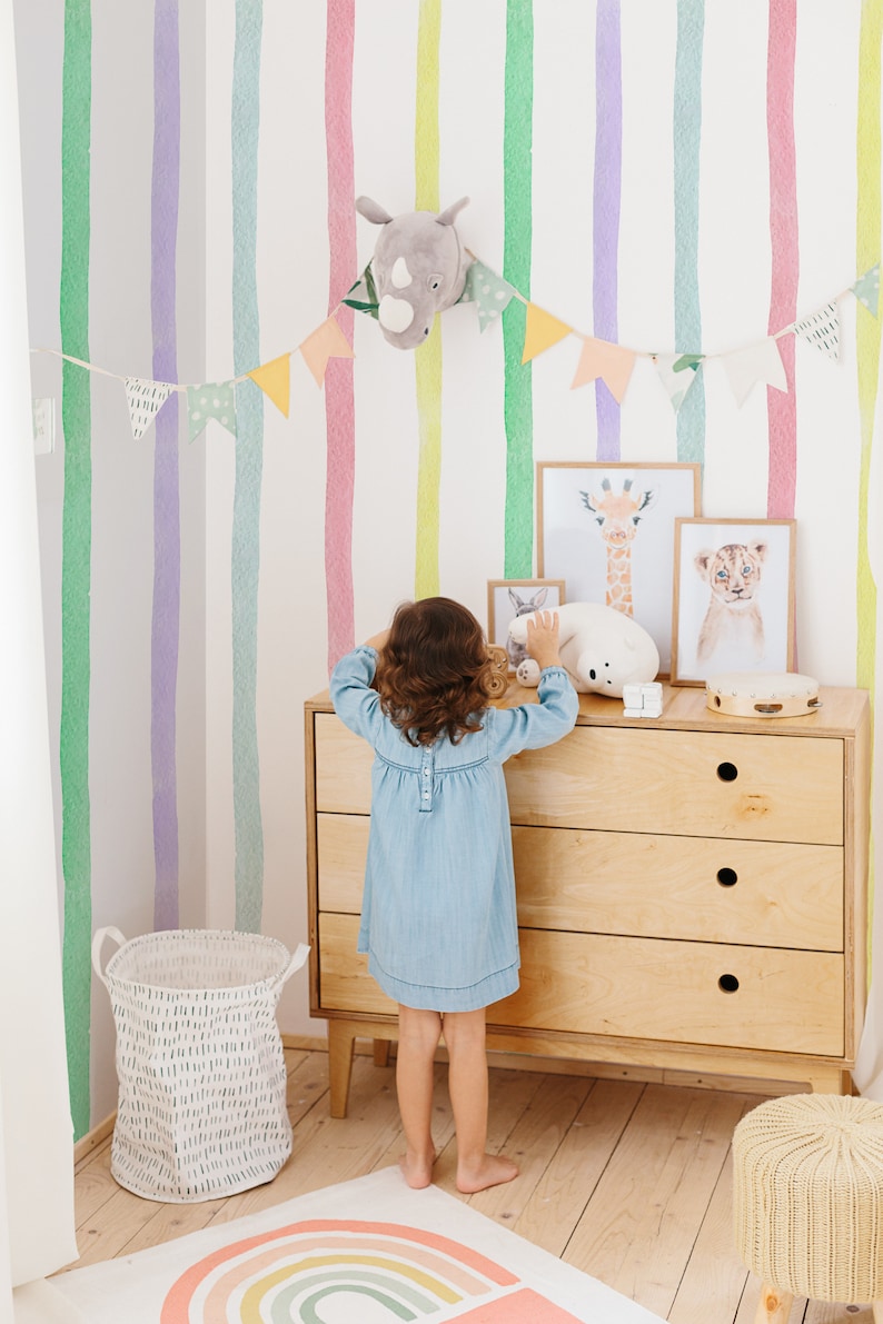K&ouml;nnte beinhalten: Ein Kind steht vor einer Kommode mit einem wei&szlig;en Hintergrund, der mit bunten Aquarellstreifen verziert ist. Die Kommode hat drei Schubladen und ist aus hellem Holz. &Uuml;ber der Kommode befinden sich Tierbilder, darunter eine Giraffe, ein L&ouml;we und ein Kaninchen. Ein wei&szlig;es Stofftier liegt auf der Kommode. Ein Regenbogen-Teppich liegt auf dem Boden vor der Kommode.