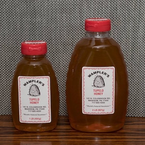 May include: Two glass bottles of WAMPLER'S Tupelo Honey. The smaller bottle is 1 lb (454g) and the larger bottle is 2 lb (907g). Both bottles have red lids and are filled with a golden honey.