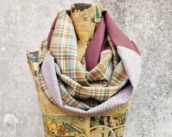 May include: A plaid infinity scarf in shades of green, blue, and tan, with a solid burgundy accent. The scarf is draped over a mannequin covered in vintage comic book pages, set against a textured gray wall.