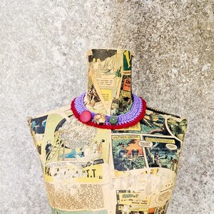 May include: A purple and red crocheted necklace with a red button and a green button on a vintage paper mache mannequin.