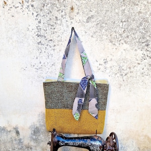 una borsa da spalla in due tessuti, una tela verde e bianca e un tessuto giallo ocra, con pochette interna, manici e piccolo fiocco in cotone africano verde, beige e blu. La borsa è appesa a un muro grigio, sopra una vecchia macchina da cucire