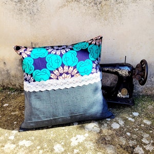 May include: A decorative cushion with a teal, blue and white patterned fabric. The cushion has a white lace trim and a grey fabric backing. The cushion is sitting on a concrete surface next to an old sewing machine.