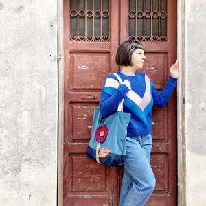 May include: A woman wearing a blue, pink and white striped jumper, blue jeans and white trainers is standing in front of a red wooden door. She is holding a blue tote bag with a flower design on the front.