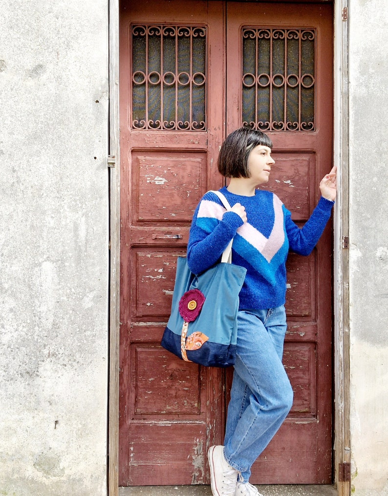 May include: A woman wearing a blue, pink and white striped jumper, blue jeans and white trainers is standing in front of a red wooden door. She is holding a blue tote bag with a flower design on the front.