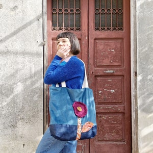 May include: A woman stands in front of a wooden door, holding a blue tote bag with a pink flower design. The bag has a brown button in the centre of the flower.