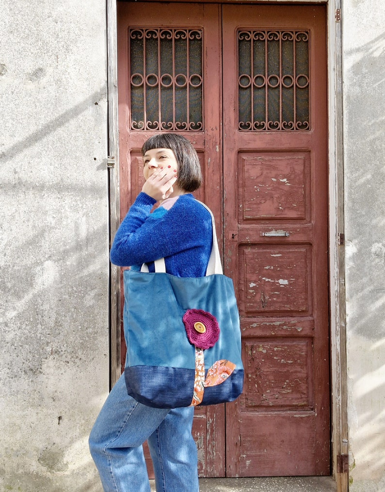 May include: A woman stands in front of a wooden door, holding a blue tote bag with a pink flower design. The bag has a brown button in the centre of the flower.