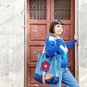 May include: A blue and denim tote bag with a pink flower applique and a brown stem. The bag is being held by a person wearing a blue and white striped sweater and blue jeans.