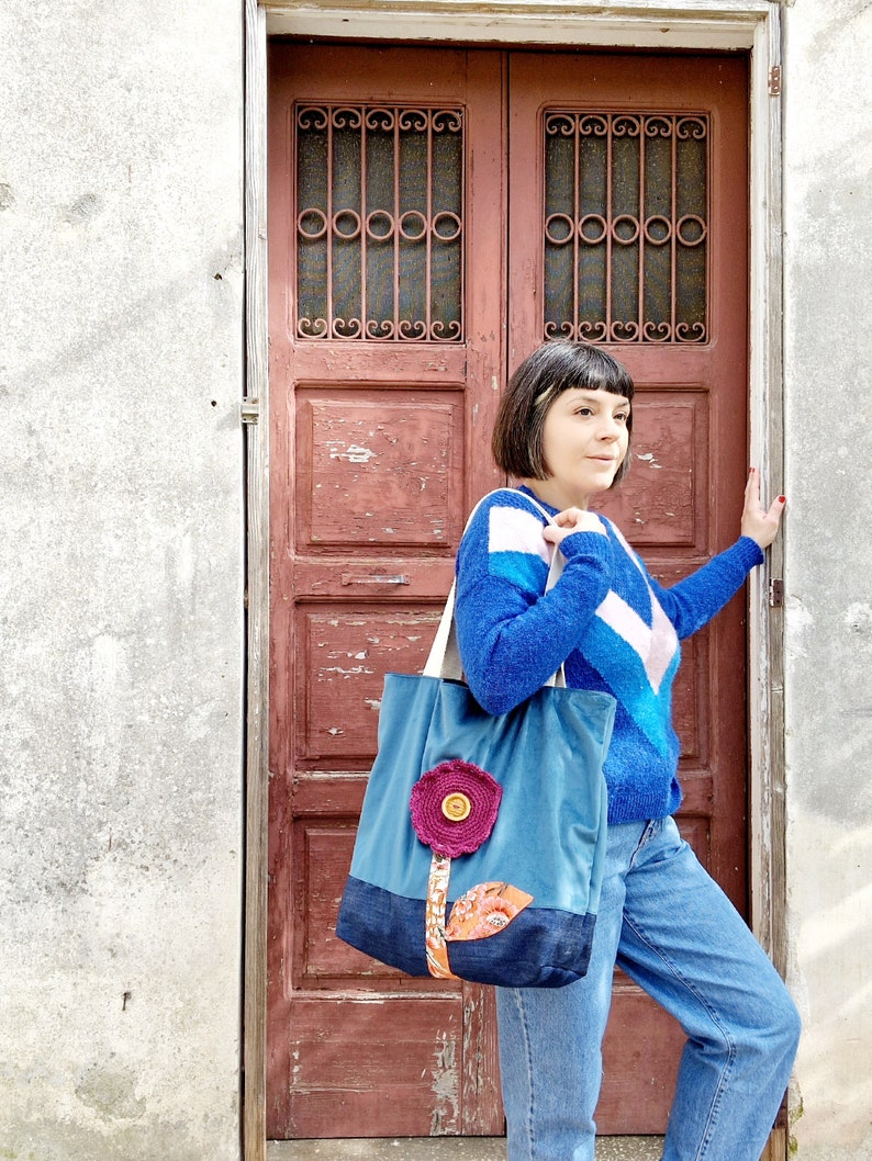 May include: A blue and denim tote bag with a pink flower applique and a brown stem. The bag is being held by a person wearing a blue and white striped sweater and blue jeans.