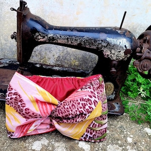 May include: A pink, yellow and brown patterned fabric headband tied in a bow. The headband is sitting on top of an old, black sewing machine.