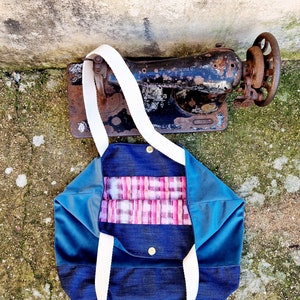 May include: A blue and denim tote bag with white straps and a pink and white plaid lining. The bag is lying on a concrete surface.