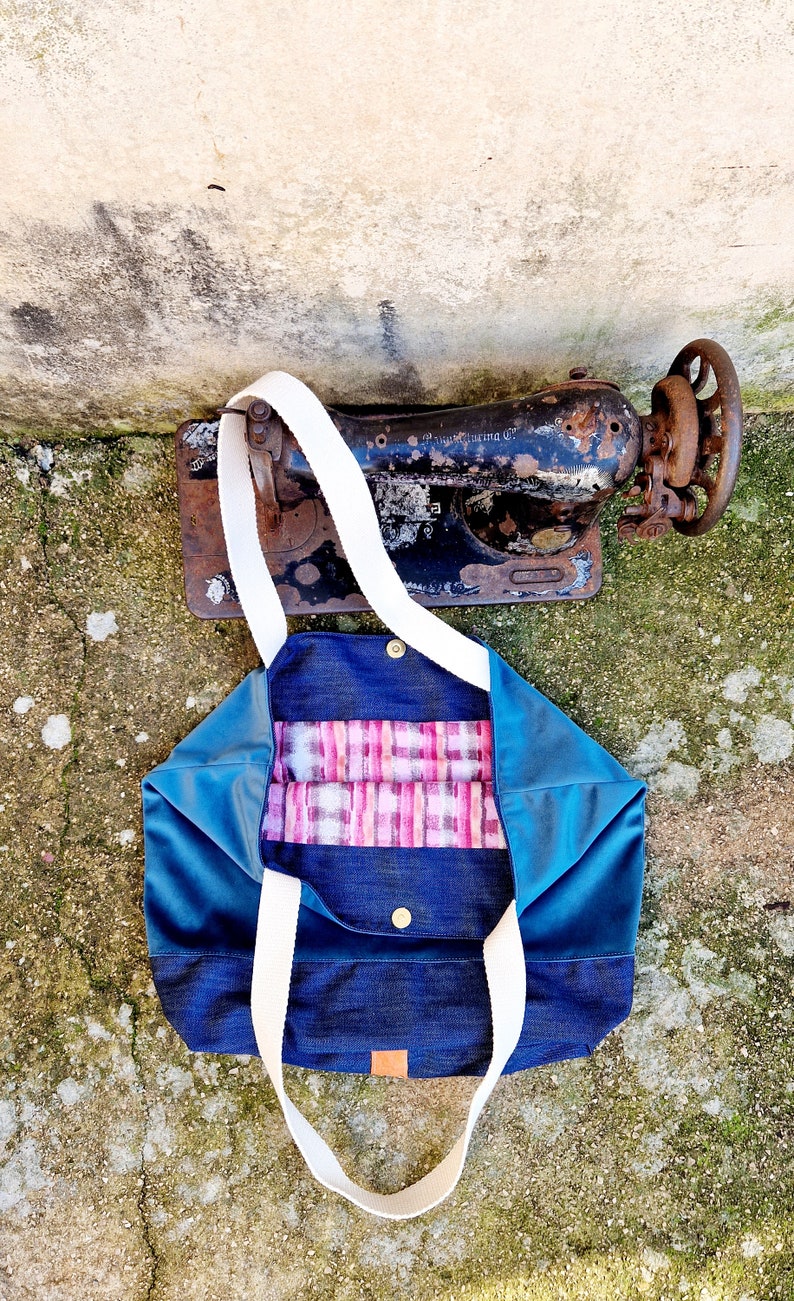 May include: A blue and denim tote bag with white straps and a pink and white plaid lining. The bag is lying on a concrete surface.