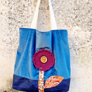 May include: A blue tote bag with a denim base and a crocheted flower appliqu&eacute;. The flower is pink with a brown button in the centre. The bag has white straps and is sitting on top of an old sewing machine.