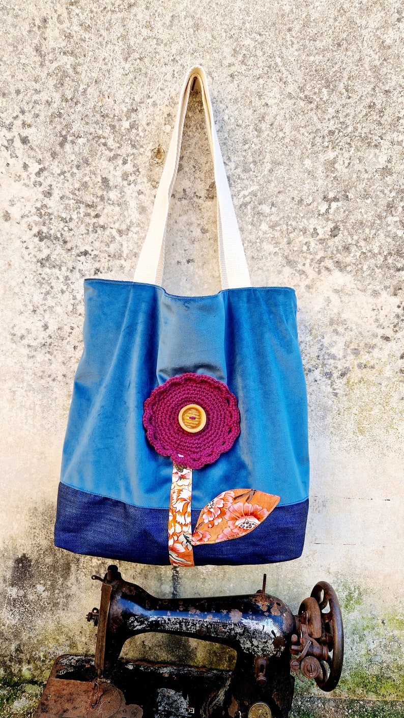 May include: A blue tote bag with a denim base and a crocheted flower appliqu&eacute;. The flower is pink with a brown button in the centre. The bag has white straps and is sitting on top of an old sewing machine.