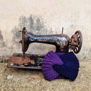 Puede incluir: Una diadema tejida a mano, en tonos morado y azul marino, junto a una máquina de coser negra vintage. La diadema presenta un diseño de nudo retorcido. El fondo es una pared texturizada de tonos neutros y una superficie de hormigón.