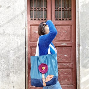 May include: A woman wearing a blue jumper and jeans is carrying a blue and denim tote bag with a pink flower design. The bag has a large button in the centre of the flower.