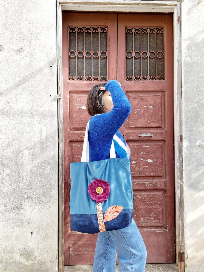 May include: A woman wearing a blue jumper and jeans is carrying a blue and denim tote bag with a pink flower design. The bag has a large button in the centre of the flower.