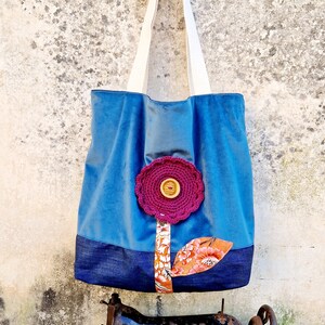 May include: A blue tote bag with a crocheted flower design and a denim bottom. The bag is hanging on a wall and there is an old sewing machine in the foreground.
