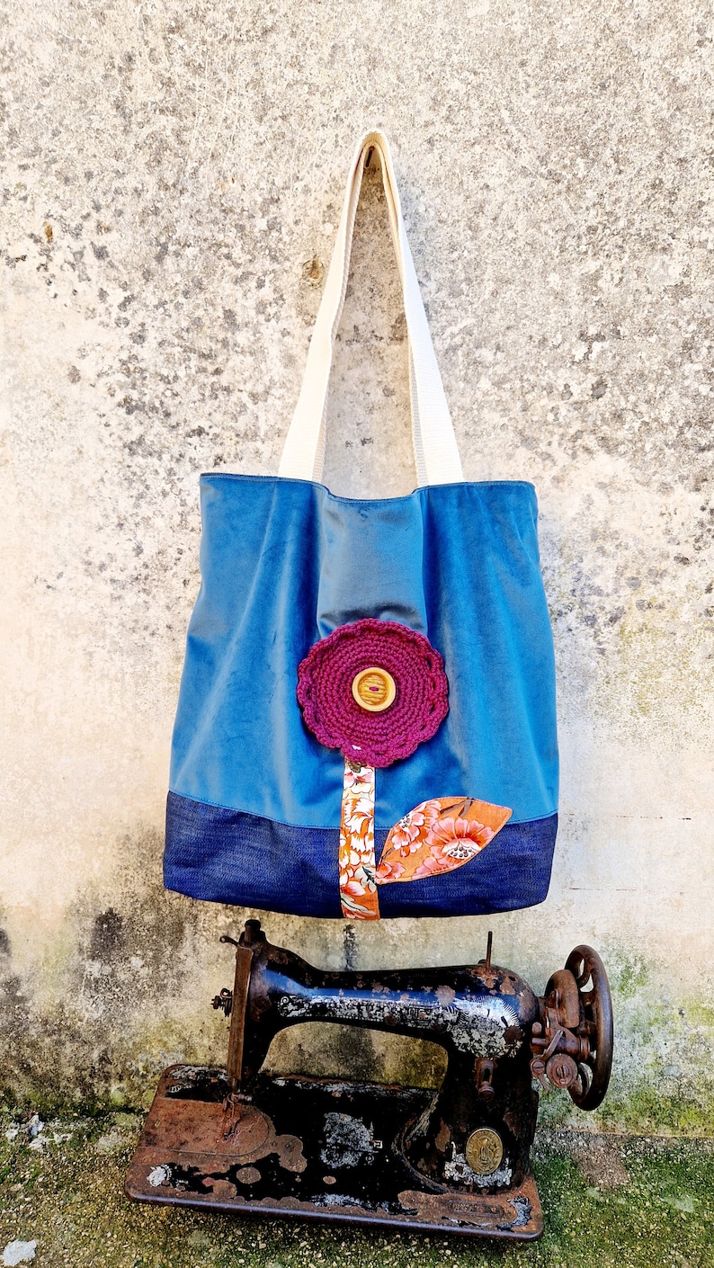 May include: A blue tote bag with a crocheted flower design and a denim bottom. The bag is hanging on a wall and there is an old sewing machine in the foreground.