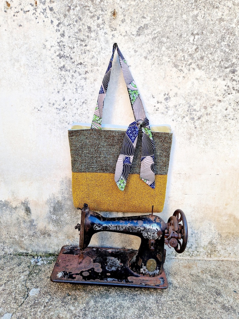una borsa da spalla in due tessuti, una tela verde e bianca e un tessuto giallo ocra, con pochette interna, manici e piccolo fiocco in cotone africano verde, beige e blu. La borsa è appesa a un muro grigio, sopra una vecchia macchina da cucire