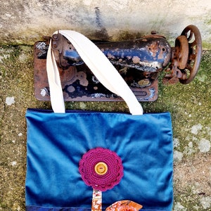 May include: A blue tote bag with a denim base and a pink crocheted flower with a wooden button in the centre. The flower is attached to a brown fabric stem with a floral print leaf.