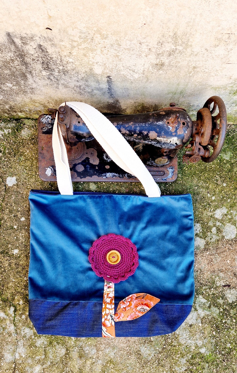May include: A blue tote bag with a denim base and a pink crocheted flower with a wooden button in the centre. The flower is attached to a brown fabric stem with a floral print leaf.
