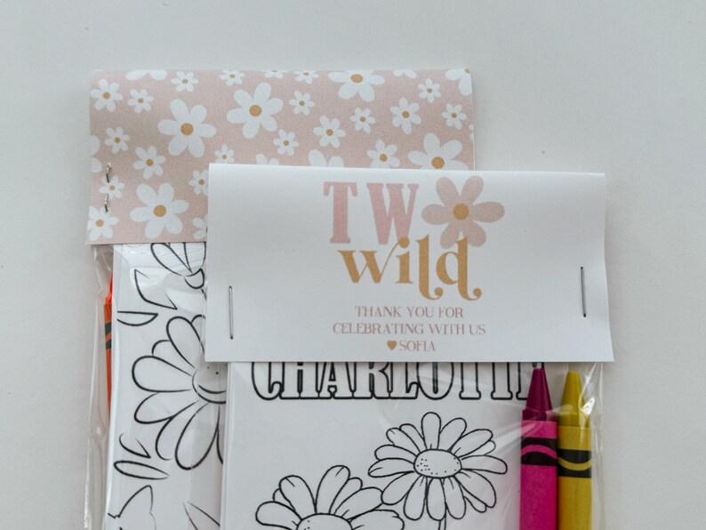 May include: A pink and white favor bag with a white card that says "Two Wild" in pink and gold letters. The card also says "Thank you for celebrating with us" and "Sofia" in pink letters. The bag contains a coloring page with a daisy design and three crayons.