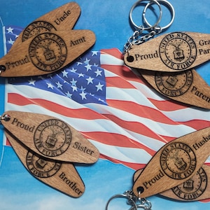 Peut inclure: Porte-clés en bois avec le logo de l'US Air Force et les mots "Proud", "Uncle", "Aunt", "Grand Parents", "Parent", "Sister", "Brother", "Husband", et "Wife". Les porte-clés sont affichés sur un drapeau américain rouge, blanc et bleu.