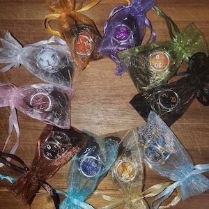 Puede incluir: Una colección de dados de colores, cada uno dentro de una bolsa de organza transparente con una cinta a juego. Los dados presentan varios colores y números, perfectos para juegos de mesa. Las bolsas están dispuestas en un patrón circular sobre una superficie de madera.