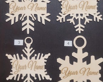 Op de afbeelding: Vier onafgewerkte houten sneeuwvlokversieringen met een gat aan de bovenkant om op te hangen. Elke versiering heeft de tekst "Your Name" gegraveerd.