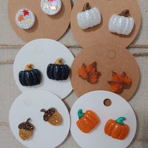 May include: Assortment of fall-themed earrings featuring pumpkins, acorns, and leaves. The earrings are made of resin and have a glittery finish.