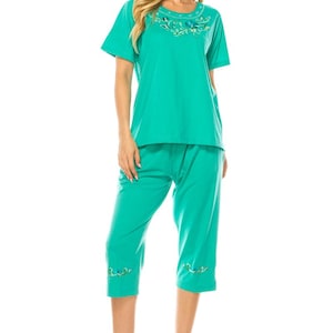 Puede incluir: Conjunto de pijama de color turquesa con una camiseta de manga corta y pantalones capri. La camiseta tiene bordados florales alrededor del cuello. Los pantalones capri tienen bordados a juego en el dobladillo. La modelo lleva sandalias transparentes de tacón.