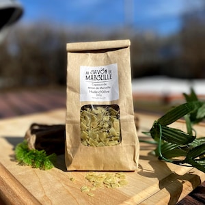 May include: A brown paper bag containing green soap flakes, with a clear window, rests on a wooden chopping board. The bag features a white label with black text: "Au Savon de Marseille". The soap flakes are scattered on the board.