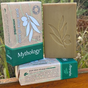 Pode incluir: Três barras de sabonete de azeite puro Mythology. Uma barra está desembrulhada, mostrando um desenho de folha. Duas barras estão em embalagens verdes e castanhas, com as palavras "pure olive oil soap unscented" e "Mythology".
