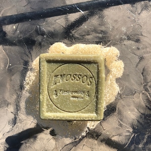 以下が含まれることがあります： 円の中に「ENOSSOS」と「First quality」の文字が書かれた緑色の石鹸。石鹸は自然なロوفスポンジの上に置かれています。