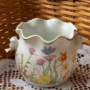 May include: A white ceramic bowl with a ruffled rim and two side handles. The exterior features a colorful floral pattern with pink, blue, orange, and yellow flowers on green stems. The rim is accented with a thin green line around the edge.
