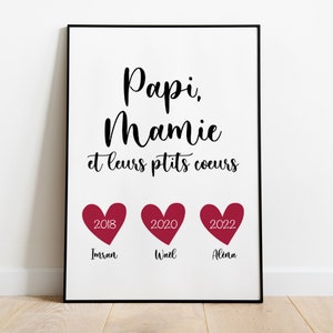 May include: A white framed print with black text that reads "Papi, Mamie et leurs ptits coeurs". Three red hearts with the years 2018, 2020, and 2022 are below the text. The names Imran, Wael, and Alena are written below the hearts.